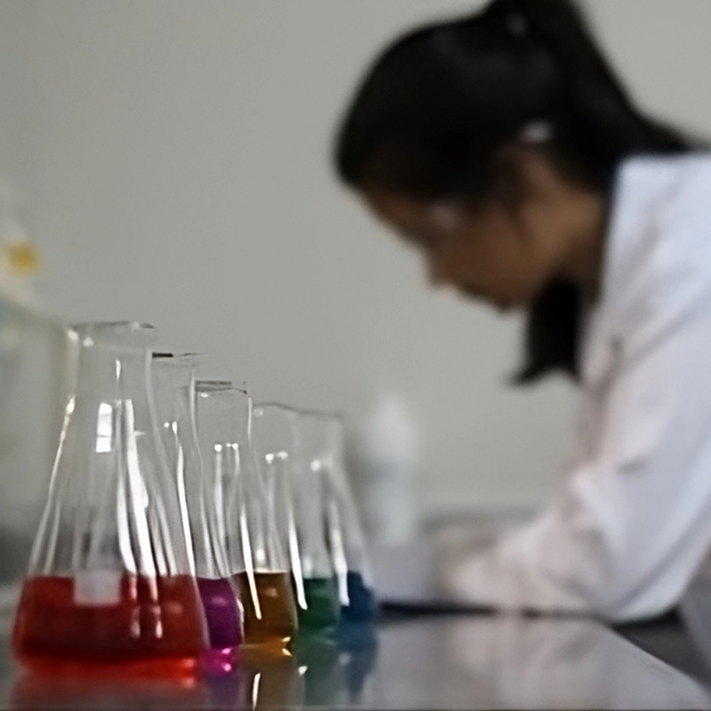 Bancada de laboratório com frascos contendo líquidos coloridos em destaque e uma pessoa desfocada ao fundo.